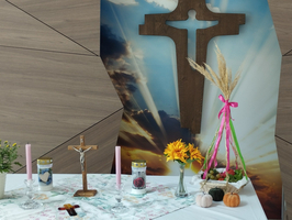 Ein Tisch mit einem Holzkreuz, Blumen und Kerzen vor einer bunten Hintergrundgestaltung.