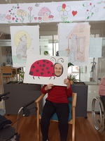 Eine Person sitzt in einem Stuhl und hält ein großes Bild eines Marienkäfers vor ihr Gesicht. Im Hintergrund sind bunte Zeichnungen von Tieren und Blumen an der Wand zu sehen. Die Umgebung wirkt freundlich und einladend.