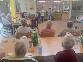 Eine Gruppe von älteren Menschen sitzt an Tischen in einem Gemeinschaftsraum. Eine Frau steht vorne und spricht, während andere zuhören. Im Hintergrund sind Getränke und Snacks auf dem Tisch sichtbar. An der Wand hängen Dekorationen mit dem Wort „Zusammenhalt“.