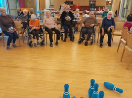 Ältere Menschen sitzen in Rollstühlen und spielen mit blauen Bowlingkegeln.