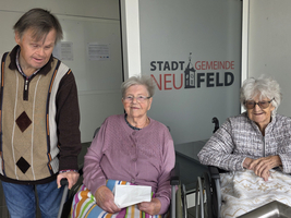 Ein Mann steht neben zwei älteren Frauen in Rollstühlen, lächelnd.