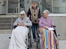 Drei Personen im Rollstuhl vor einem Gebäude, eine Person steht hinter ihnen.