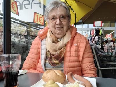 Eine ältere Frau sitzt am Tisch mit einem Teller voller Würstchen und Brötchen.