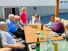 Eine Gruppe älterer Menschen sitzt an einem Tisch im Freien. Sie unterhalten sich und genießen Snacks und Getränke. Die Szene vermittelt eine freundliche und gesellige Stimmung.