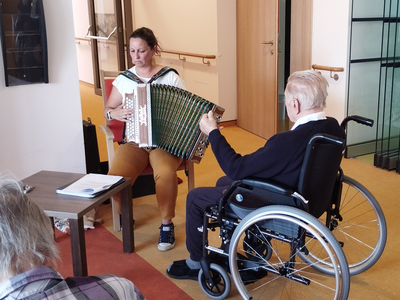 Eine Frau spielt Akkordeon, während ein älterer Mann im Rollstuhl zuhört.