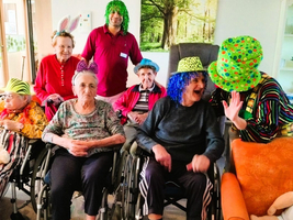 Ältere Menschen in Rollstühlen feiern, tragen bunte Hüte und Perücken.