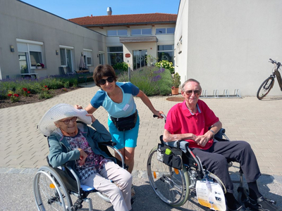 Eine Frau schiebt zwei ältere Menschen in Rollstühlen an einem sonnigen Tag.