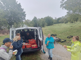 Zu sehen ist ein offener Lieferwagen mit vielen Obstkisten. Davor sieht man 7 Menschen bei der Jause. 