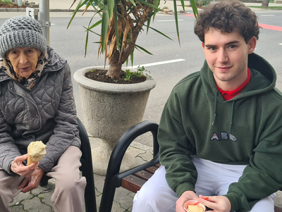 Zwischen zwei Personen, die Eis essen, sitzt eine ältere Dame und ein junger Mann.