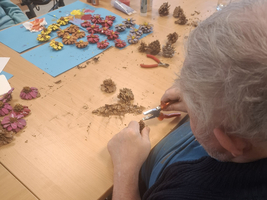 Ein älterer Mann arbeitet an einem Tisch, umgeben von Bastelmaterialien wie Tannenzapfen und getrockneten Blumen. Er schneidet mit einer Schere oder Zange, während andere Materialien auf dem Tisch verteilt sind. Im Hintergrund sind weitere Personen und Bastelutensilien sichtbar.