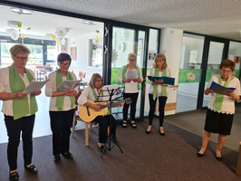 Sechs Frauen in Blusen mit grünen Schals stehen in einem Raum und singen. Eine Frau sitzt mit einer Gitarre und spielt dazu. Alle halten Notenblätter und scheinen bei ihrem Auftritt konzentriert. Im Hintergrund sind helle Möbel und Dekoration sichtbar.