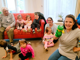 Eine Gruppe von Kindern sitzt auf einem roten Sofa, während eine Frau mit einem Handpuppenfrosch lächelt. Eine ältere Dame beobachtet die Szene. Die Kinder sind fröhlich und in bunten Kleidern, und es herrscht eine freundliche, familienorientierte Atmosphäre.