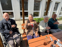 Vier ältere Menschen sitzen an einem Tisch im Freien, genießen den Sonnenschein und heben Gläser in einer geselligen Runde. Im Hintergrund sind Pflanzen und ein Gebäude zu sehen.