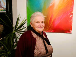 Ältere Frau mit grauen Haaren, lächelnd vor einem bunten Kunstwerk.