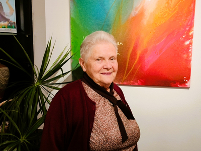 Ältere Frau mit grauen Haaren, lächelnd vor einem bunten Kunstwerk.