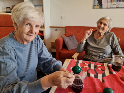 Zwei ältere Frauen sitzen an einem Tisch mit einem roten Tuch. Eine Frau hält einen Krug mit einer dunklen Flüssigkeit, während die andere lächelt und einen Finger hebt. Vor ihnen stehen Gläser mit Getränken. Im Hintergrund sieht man eine gemütliche Wohnumgebung.