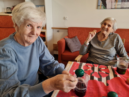 Zwei ältere Frauen sitzen an einem Tisch mit einem roten Tuch. Eine Frau hält einen Krug mit einer dunklen Flüssigkeit, während die andere lächelt und einen Finger hebt. Vor ihnen stehen Gläser mit Getränken. Im Hintergrund sieht man eine gemütliche Wohnumgebung.