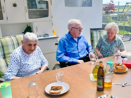 Drei ältere Personen sitzen an einem Tisch im Freien, umgeben von Essen und Getränken. Zwei Frauen, eine mit grauem Haar und eine mit weißen Haaren, und ein Mann in einem blauen Hemd, scheinen sich zu unterhalten. Die Atmosphäre ist freundlich und entspannt.