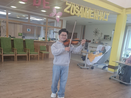 Ein Junge steht in einem Raum und spielt Geige. Im Hintergrund sind Stühle und eine Sitzecke zu sehen. An der Wand hängt ein dekoratives Banner mit dem Wort 