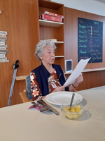 Eine ältere Frau liest ein Dokument am Tisch, vor ihr ein Teller mit Obst.