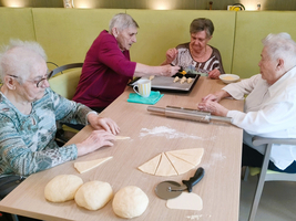 Vier ältere Frauen sitzen an einem Tisch und backen. Zwei von ihnen kneten Teig, während die anderen mit einer Backform und Teigstücken arbeiten. Auf dem Tisch liegen Teigstücke und eine Backrolle. Die Atmosphäre ist freundlich und aktiv.