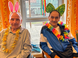 Zwei Männer tragen bunte Kostüme und feiern mit Accessoires am Fenster.