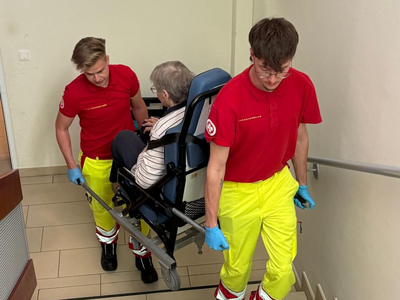 Zwei Sanitäter in roten Shirts und gelben Hosen tragen eine tragbare Trage mit einer Person die auf ihr sitzt, die Treppe hinunter. Die Szene vermittelt eine Notfallbergungssituation.