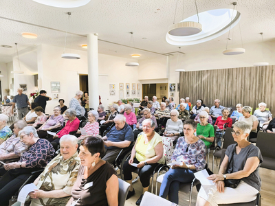 Eine Gruppe älterer Menschen sitzt in einemhellen Raum und hört aufmerksam zu.