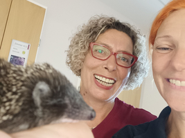 Eine Frau mit lockigem Haar und Brille hält einen Igel in der Hand, während eine andere Frau mit orangefarbenen Haaren neben ihr steht. Beide lächeln fröhlich in die Kamera. Der Hintergrund zeigt eine hell erleuchtete Inneneinrichtung.