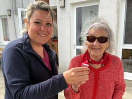 Eine lächelnde Pflegekraft steht neben einer älteren Frau, die Sonnenbrille trägt und einen roten Pullover anhat. Die Frau hält einen kleinen Becher, während die Pflegekraft ihr freundlich zuprostet. Im Hintergrund sind helle Gebäudewände und ein gepflasterter Boden sichtbar.