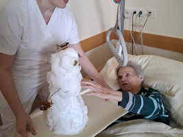 Eine Pflegerin zeigt einer älteren Frau einen Schneemann im Bett.